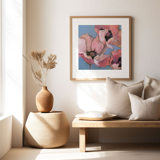 Framed print of pink poppies on a blue background in a brightly lit modern room with a wooden vase and dried flowers and a bench with natural color pillows.