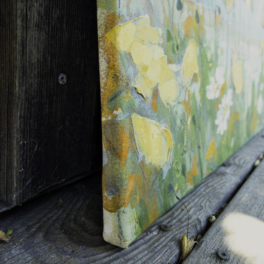 A close-up of a textured canvas painting leaning against a dark wooden surface. The artwork features a floral design with prominent yellow and white blossoms on a green and gray background.