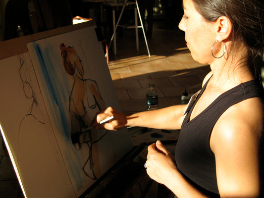A woman in a black tank top paints a watercolor portrait of a standing figure on an easel. Sunlight highlights her focused expression and brushstrokes.