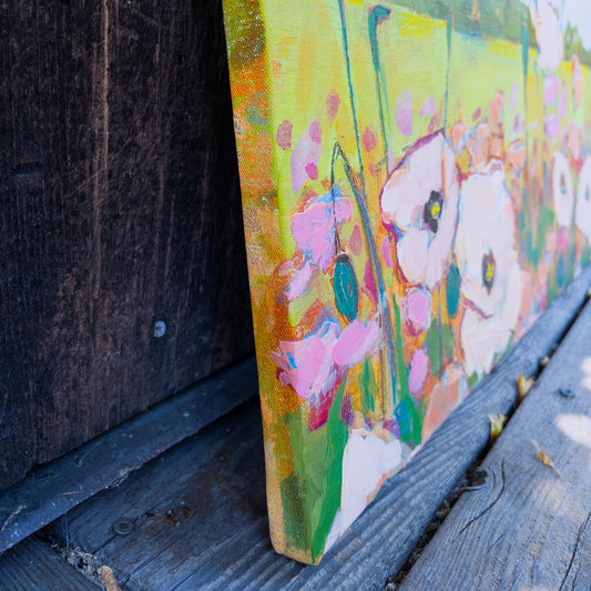 A close-up of a colorful floral canvas painting leaning against a dark wooden surface outdoors on weathered wood planks. The artwork features large white and pink flowers with green stems and yellow-green background brushstrokes.